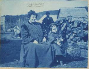 First white child born in Cook Inlet, Martha White, and her mother, left, in Sunrise, Alaska in 1898. The child is wearing a hat and holding a cat. A man, several wooden buildings and a pile of logs are visible in the background. Photograph taken during the 1898 Cook's Inlet Exploring Expedition led by Edwin F. Glenn on behalf of the U.S. Army. The photographer is unidentified. Title taken from caption written on back of photograph. [Edwin F. Glenn papers, Archives and Special Collections, Consortium Library, University of Alaska Anchorage] 